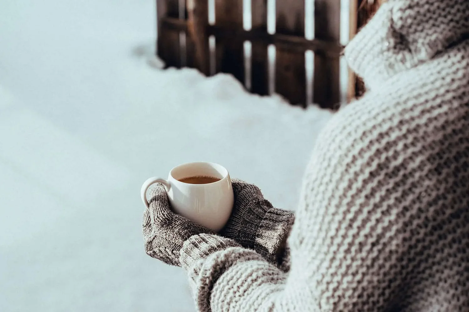 Eine Person trägt einen warmen, gestrickten Pullover und graue Handschuhe. Sie hält eine Tasse Kaffee oder Tee in den Händen. Im Hintergrund liegt Schnee, was auf eine kalte Umgebung hinweist.