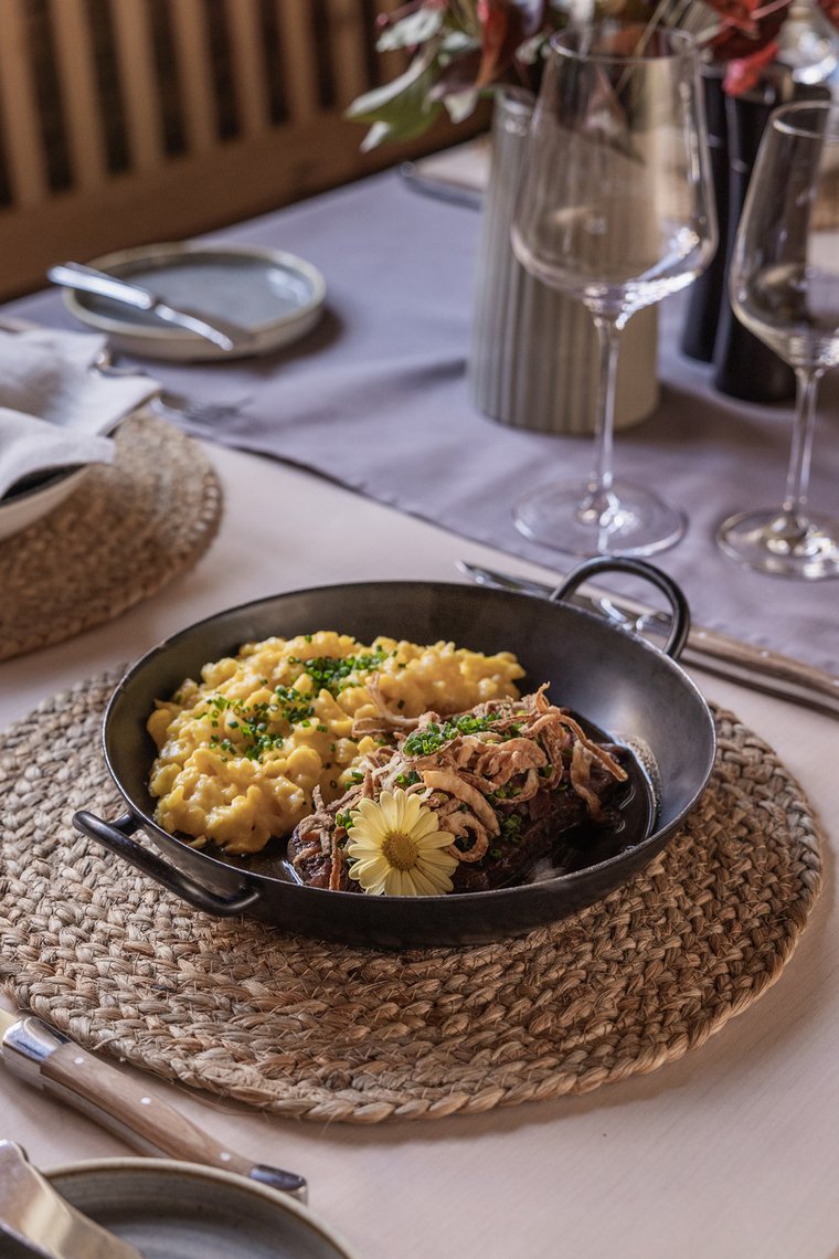 Eine dunkle Schüssel auf einem runden Tisch, gefüllt mit cremiger Pasta und zarten Fleischstücken, garniert mit frittierten Zwiebelringen und einem essbaren Blütenblatt. Im Hintergrund sind Weingläser und dekorative Elemente zu sehen.