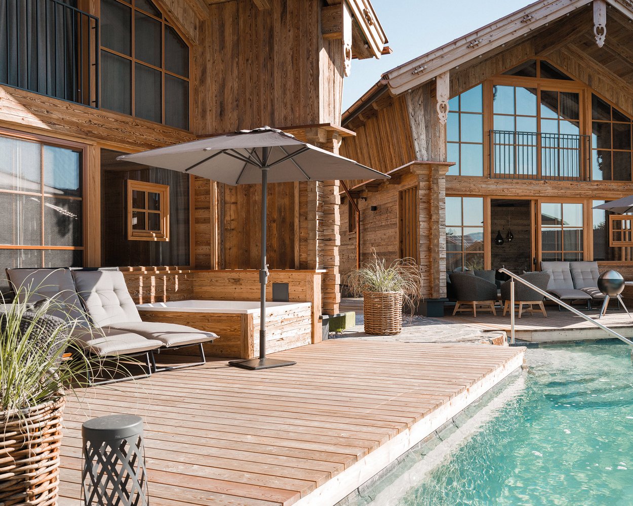 Eine moderne Holzterrasse mit einem Pool, Liegen und einem Sonnenschirm. Im Hintergrund stehen zwei große, rustikale Holzhäuser mit großen Fenstern. Grüne Pflanzen sind um die Terrasse herum platziert, und die Sonne scheint auf die Szene.
