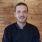 A smiling chef wearing a black culinary jacket stands against a wooden background. He appears approachable and confident, suggesting expertise in cooking.