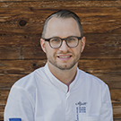 A smiling chef wearing a white chef's jacket and glasses poses in front of a rustic wooden background. The chef appears approachable and professional, embodying a culinary context.