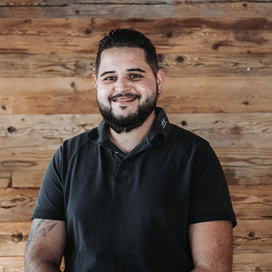 A smiling man with a beard is seated in front of a wooden backdrop. He is wearing a black polo shirt and has tattoos visible on his forearm. The image conveys a friendly and approachable demeanor.
