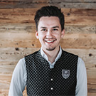 Ein junger Mann mit einem freundlichen Lächeln trägt eine elegante Weste über einem hellen Hemd. Er steht vor einer rustikalen Holzverkleidung, die eine warme und einladende Atmosphäre schafft.