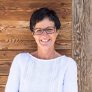 Eine lachende Frau mit kurzen, dunklen Haaren und Brille steht vor einer holzverkleideten Wand. Sie trägt ein weißes Oberteil und hat eine freundliche Ausstrahlung.