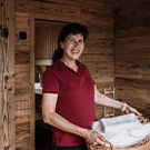 Eine lächelnde Frau in einem roten Poloshirt steht in einem Holzraum und hält ein Tablett mit weißen Handtüchern. Sie wirkt freundlich und einladend. Im Hintergrund sind unscharfe Elemente eines weiteren Raumes zu erkennen.