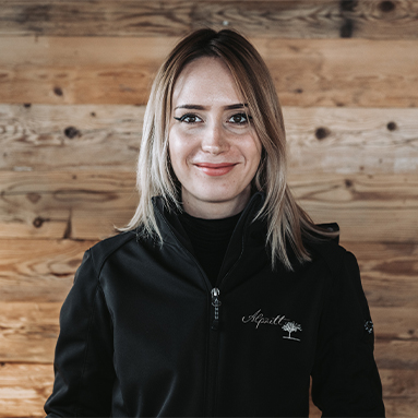 Eine lächelnde Frau mit längeren, blonden Haaren steht vor einer Holzpaneelwand. Sie trägt eine schwarze Jacke mit einem Logo auf der linken Brustseite. Ihr Gesichtsausdruck ist freundlich und einladend.
