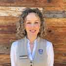 A smiling woman with curly hair is wearing a light-colored vest over a white blouse. She stands against a wooden backdrop, exuding a friendly and welcoming demeanor. A name tag is visible on her vest, suggesting she is in a professional setting.