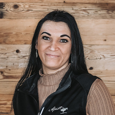 A woman with long dark hair smiles at the camera. She wears a dark vest over a light brown turtleneck. The background features wooden panels, suggesting a warm, rustic setting. Her expression conveys friendliness and approachability.