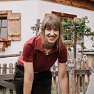 A person with medium-length hair is smiling and bending slightly forward. They are wearing a dark red polo shirt and black pants. The background features a wooden fence and greenery, suggesting an outdoor setting near a house with wooden shutters.