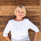 A smiling woman with short blonde hair is standing with her hands on her hips, wearing a white blouse. She is positioned in front of a rustic wooden background, conveying a friendly and approachable demeanor.