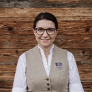 Eine Frau mit Brille in einer beigefarbenen Weste lächelt vor einer Holzwand. Sie wirkt freundlich und professionell, vermittelt ein Gefühl von Zugänglichkeit und Offenheit.