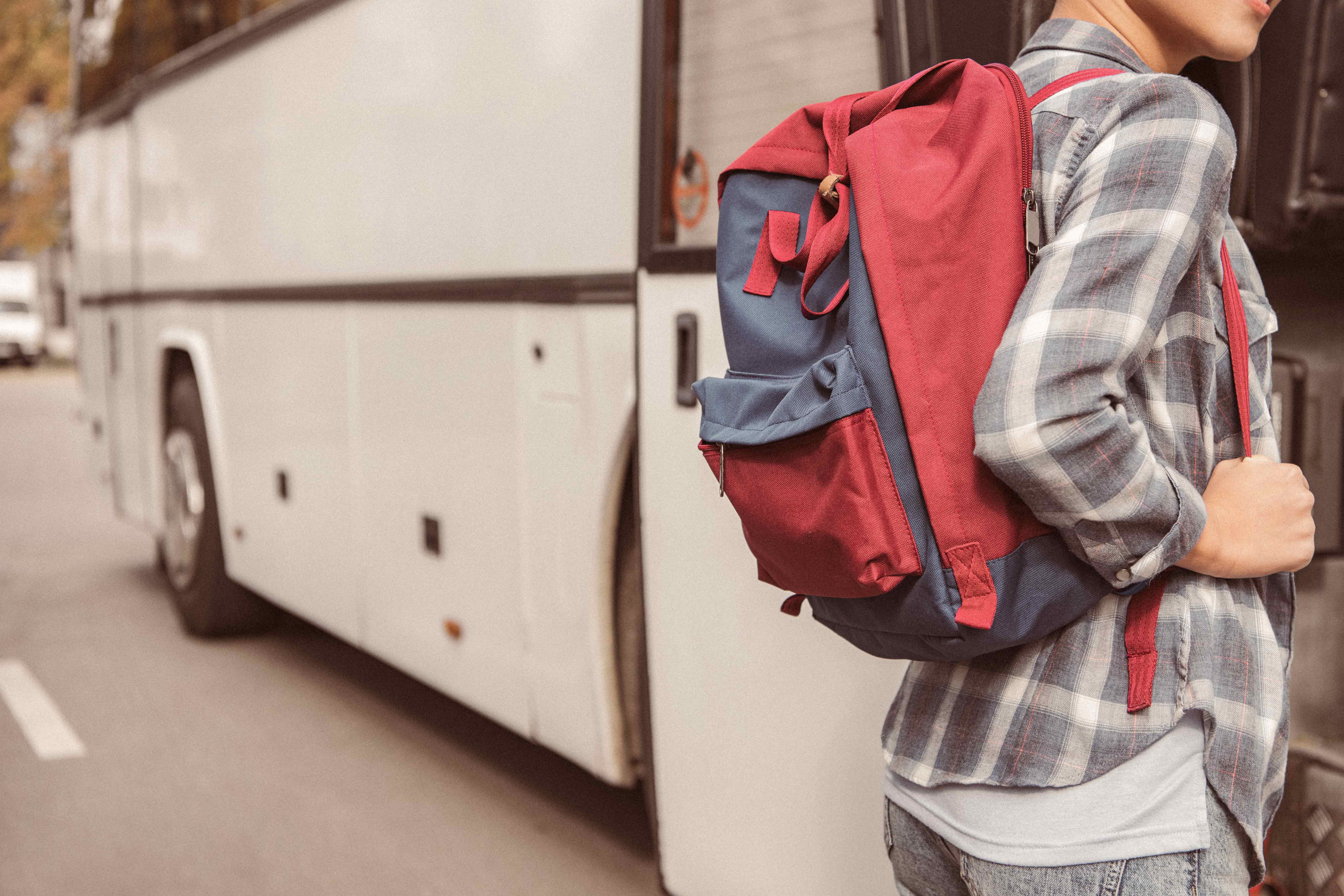 Eine Person steht an einer Haltestelle neben einem Bus und trägt einen Rucksack in rot und blau. Sie trägt ein kariertes Hemd und schaut in Richtung des Busses. Die Szene vermittelt eine wartende Stimmung.