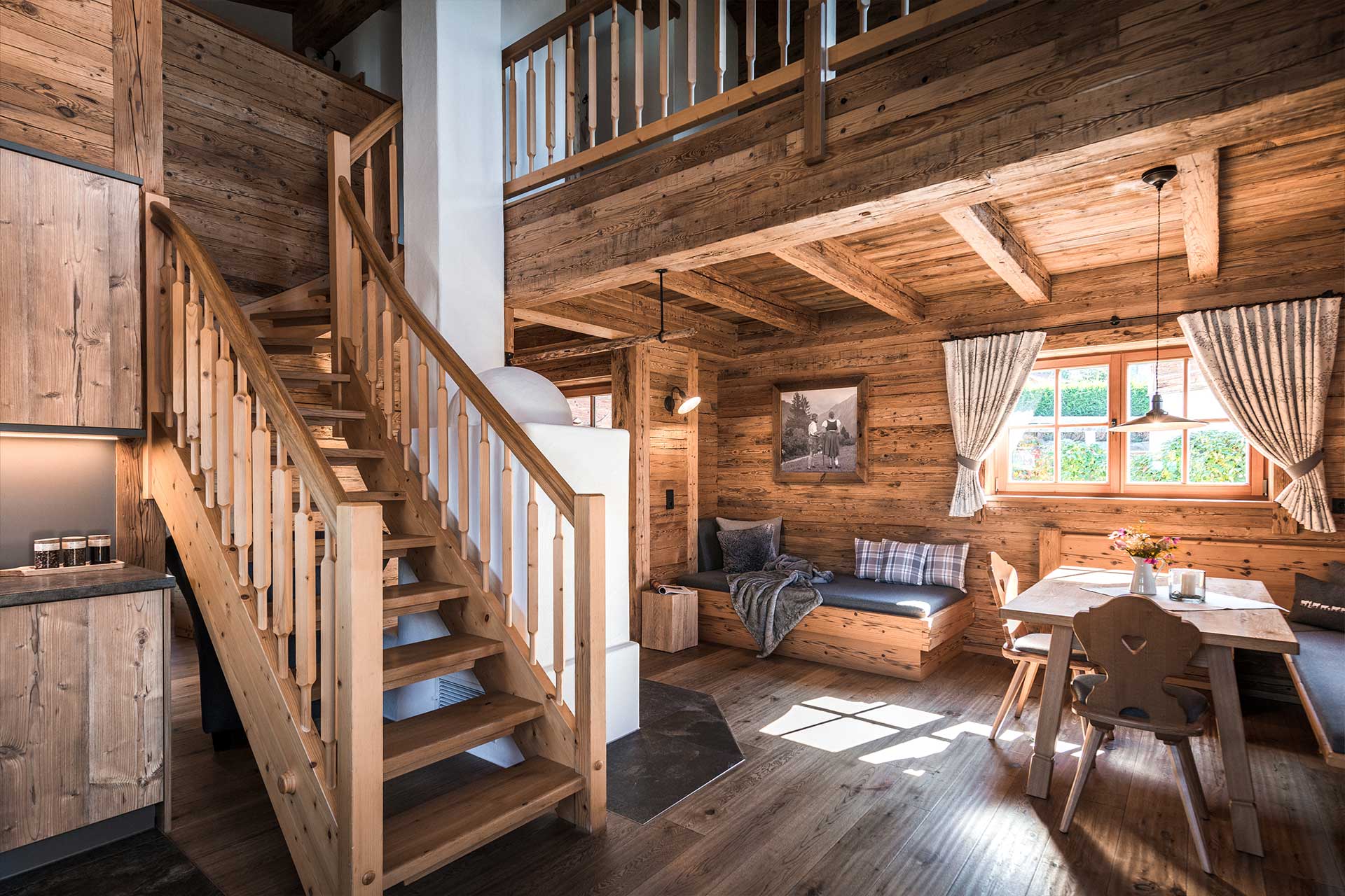 Eine helle, gemütliche Wohnstube in einem Holzhaus mit offenen Raumkonzept. Eine Treppe führt ins Obergeschoss. Links eine moderne Küchenzeile, rechts eine Sitzecke mit einem Sofa und einem Tisch, umgeben von großen Fenstern mit Vorhängen. Holzverkleidung schafft eine warme Atmosphäre.