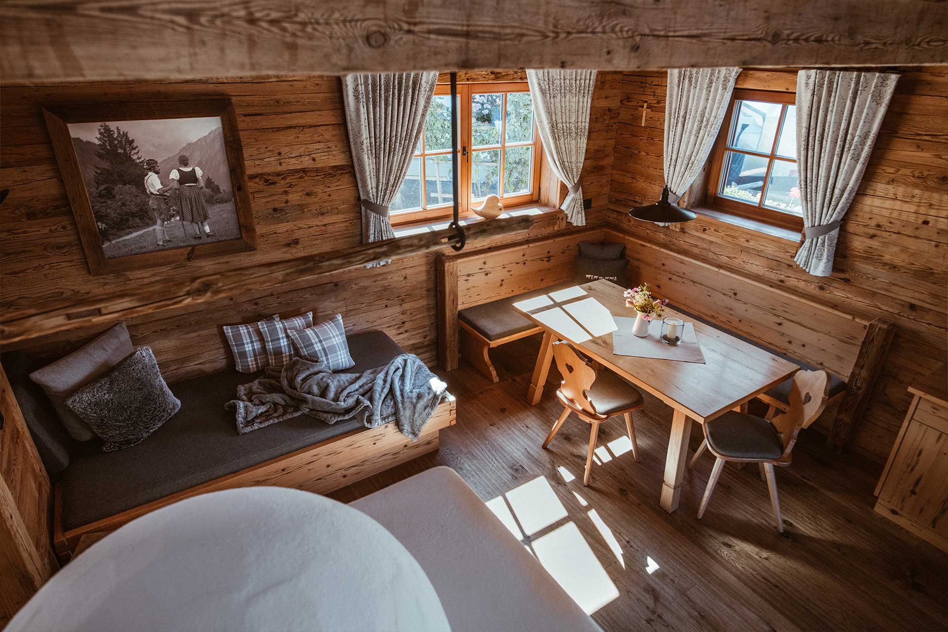 Ein gemütlicher Holzraum mit einem Sofa, einem Tisch und Stühlen. Tageslicht strömt durch zwei Fenster mit Vorhängen. Auf dem Tisch steht eine kleine Blumenvase. An der Wand hängt ein nostalgisches Bild. Der Boden ist ebenfalls aus Holz, was eine warme Atmosphäre schafft.