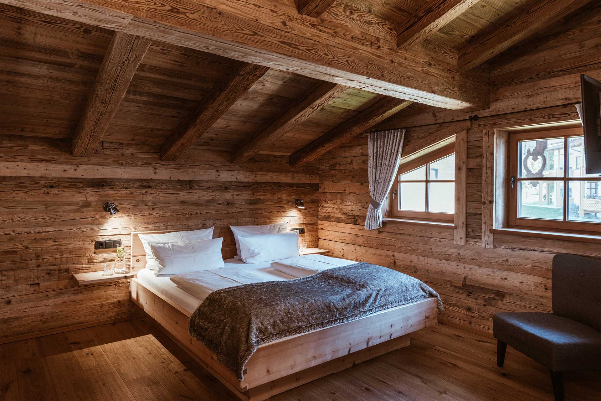 Das Bild zeigt ein gemütliches Schlafzimmer mit Holzvertäfelungen. Ein großes Bett mit weißen Bettwäsche und einer grauen Decke steht im Raum. Es gibt zwei Fenster mit Vorhängen, sowie eine bequeme Ecke mit einem Stuhl. Die Atmosphäre wirkt warm und einladend.