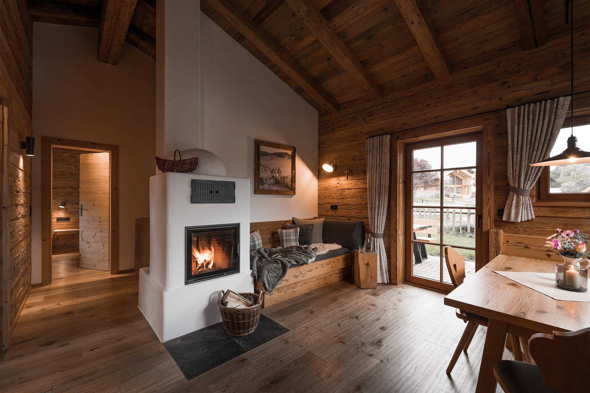 Gemütliche Holzhütte mit einem offenen Wohnbereich. Ein Kamin mit offenes Feuer steht im Vordergrund. Ein Sofa und ein Holztisch laden zum Verweilen ein. Große Fenster lassen Tageslicht herein und bieten einen Blick nach draußen. Der Raum strahlt Wärme und Ruhe aus.