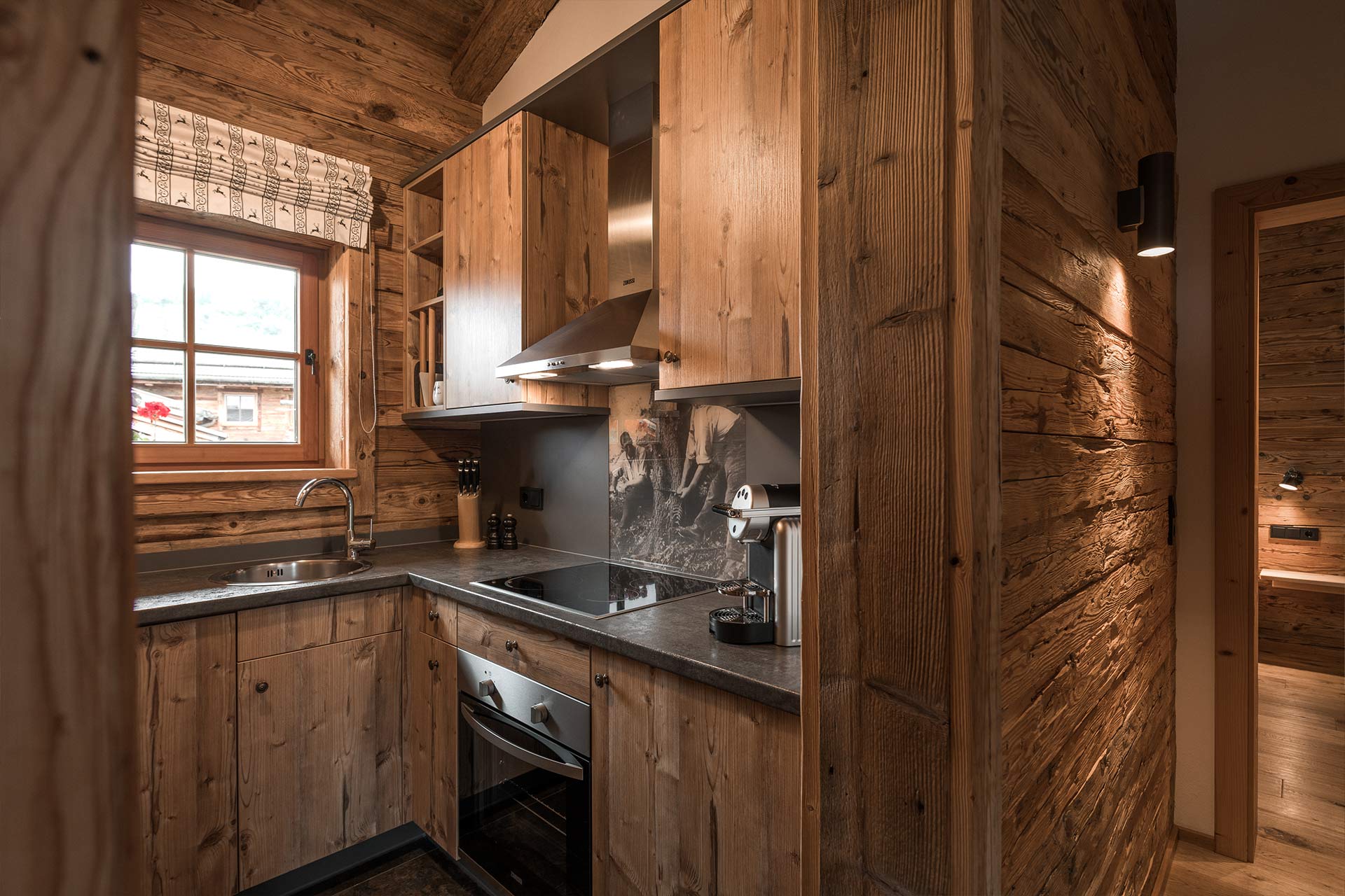 Die Küche ist aus Holz und hat eine moderne Ausstattung. Sie verfügt über eine Spüle, eine Kochplatte, einen Backofen und Küchenschränke. Ein Fenster mit einer Gardine lässt Licht herein. Die Wände sind ebenfalls holzverkleidet und schaffen eine gemütliche Atmosphäre.