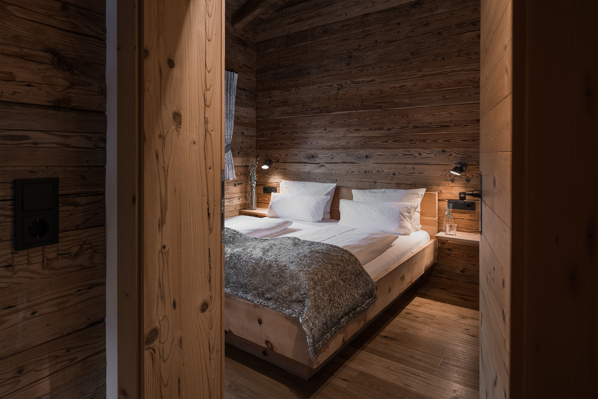 Ein gemütliches Schlafzimmer mit Holzverkleidung. Es enthält ein Doppelbett mit weißen Kissen und einer grauen Decke. An den Wänden sind Nachtlampen angebracht und ein Fenster mit Vorhängen bietet Blick nach außen. Der Boden ist aus Holz und sorgt für eine warme Atmosphäre.