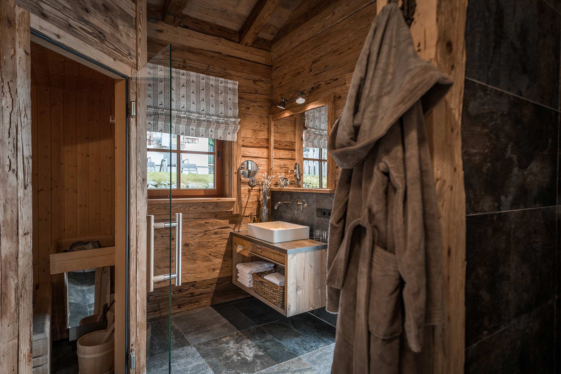 Ein modernes Bad in einem Holzhaus. Es verfügt über eine große Duschkabine, ein Waschbecken auf einem Holzschrank und einen Spiegel. Ein Handtuch hängt an einem Haken. Im Hintergrund sind Fenster mit Vorhängen und eine Holzvertäfelung zu sehen.