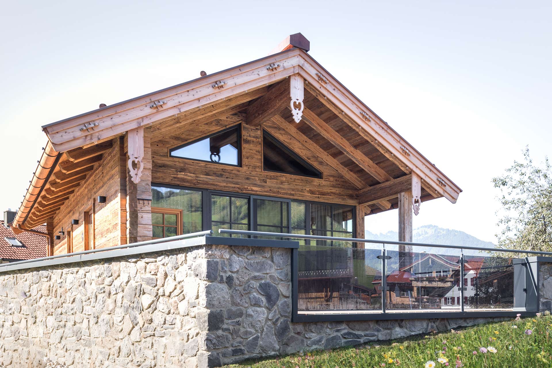 Das Bild zeigt ein modernes Holzhaus mit einem hohen Dach und großen Fenstern. Es verfügt über eine Steinmauer und ein Geländer aus Glas. Der Hintergrund zeigt eine bergige Landschaft, die dem Gebäude ein charmantes, ländliches Ambiente verleiht.