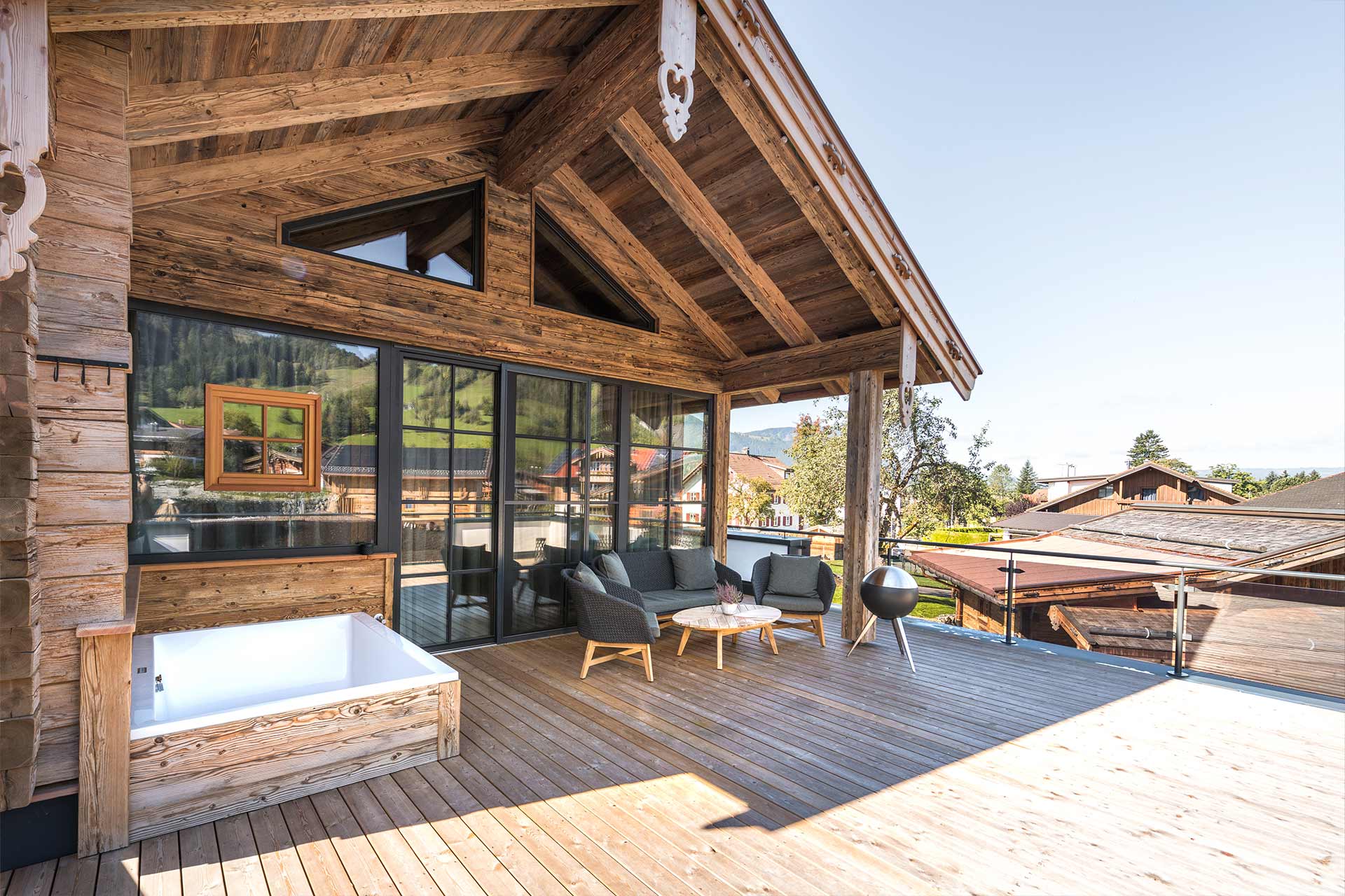 Eine moderne Holzterrasse mit entspannenden Sitzgelegenheiten und einem Holzbad. Im Hintergrund sind grüne Hügel und weitere Häuser zu sehen. Die große Glasfront lässt viel Licht herein und verbindet den Innenraum mit der Natur.