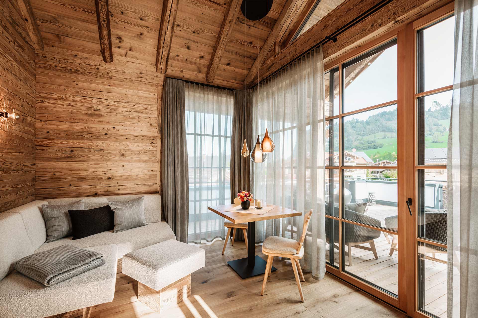 Ein gemütliches Zimmer mit Holzverkleidung. Auf einer Seite steht ein helles Sofa mit Kissen, gegenüber ein kleiner Tisch mit einer Blumendekoration. Große Fenster mit Vorhängen ermöglichen einen Blick nach draußen auf eine grüne Landschaft. Warmes Licht von modernen Lampen schafft eine einladende Atmosphäre.