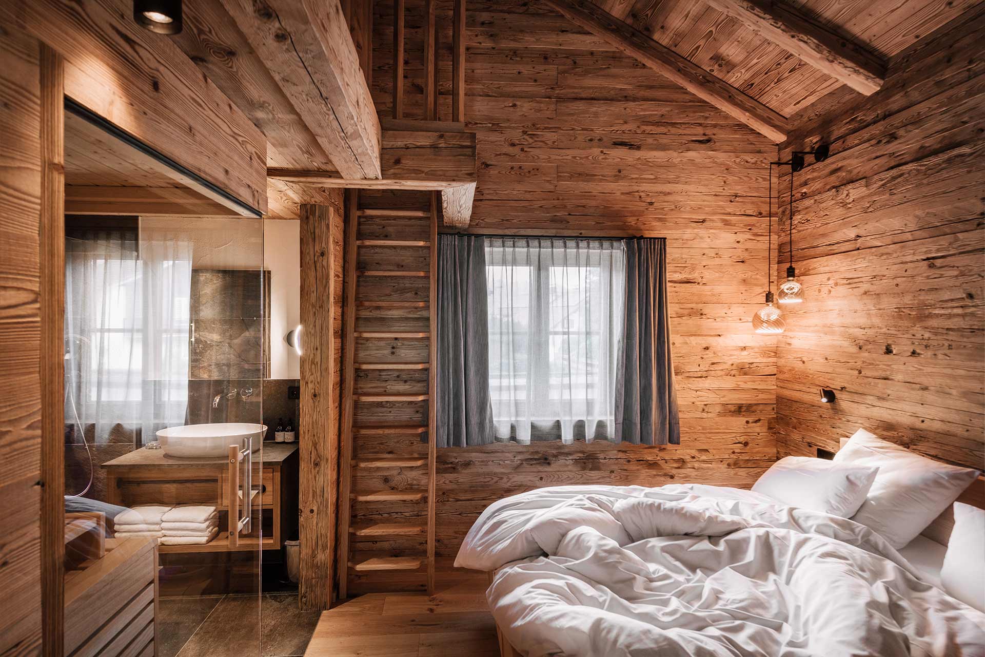 Das Bild zeigt ein gemütliches Schlafzimmer aus Holz mit einem Doppelbett und weißen Bettwäsche. Eine Treppe führt nach oben, und große Fenster lassen Tageslicht herein. Der Raum hat eine rustikale Atmosphäre, mit holzverkleideten Wänden und warmen Beleuchtungselementen.