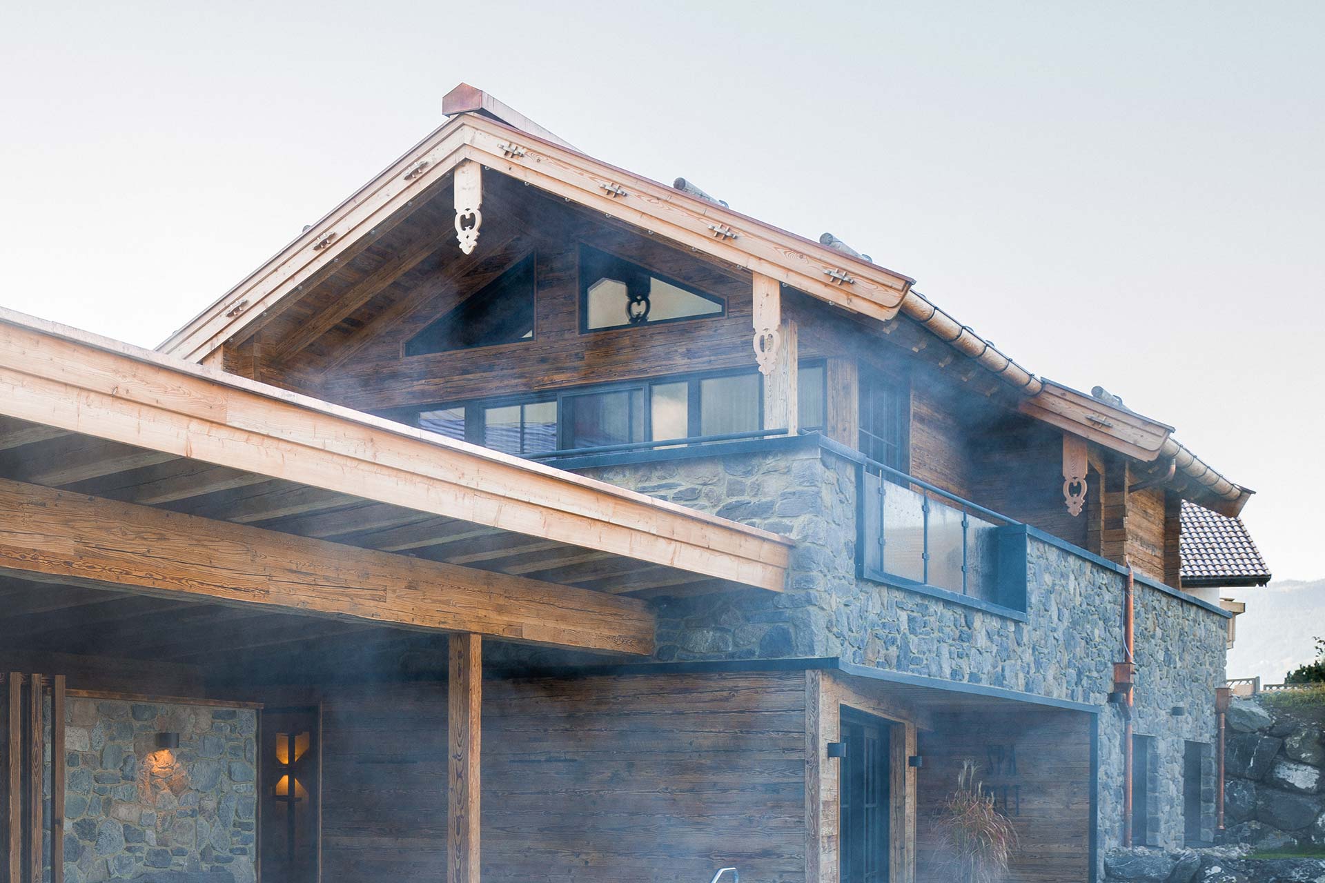 Ein modernes Haus mit einer Kombination aus Holz und Stein. Es hat große Fenster und ein leicht geneigtes Dach. Kleine ornamentale Details sind sichtbar. Im Vordergrund ist eine überdachte Terrasse zu sehen. Eine leichte Nebelbildung umgibt das Gebäude, was eine ruhige Atmosphäre schafft.