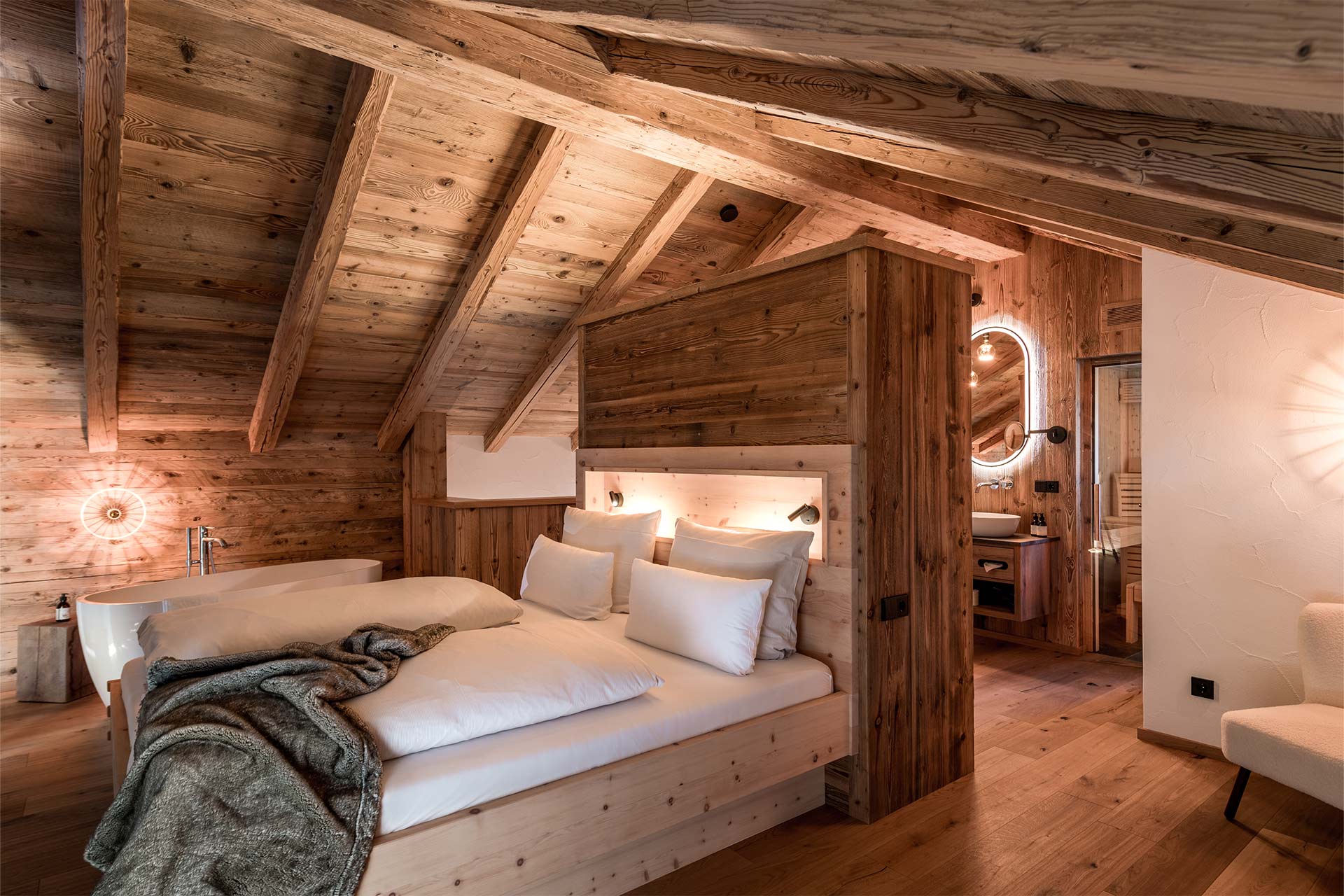 Ein modernes Schlafzimmer im alpenländischen Stil mit Holzverkleidung. Es enthält ein großes Bett mit weißen Kissen und einer Wolldecke. Die schrägen Deckenbalken und warmen Holzfarben schaffen eine gemütliche Atmosphäre. Ein Badezimmerbereich mit sanitären Einrichtungen ist im Hintergrund sichtbar.