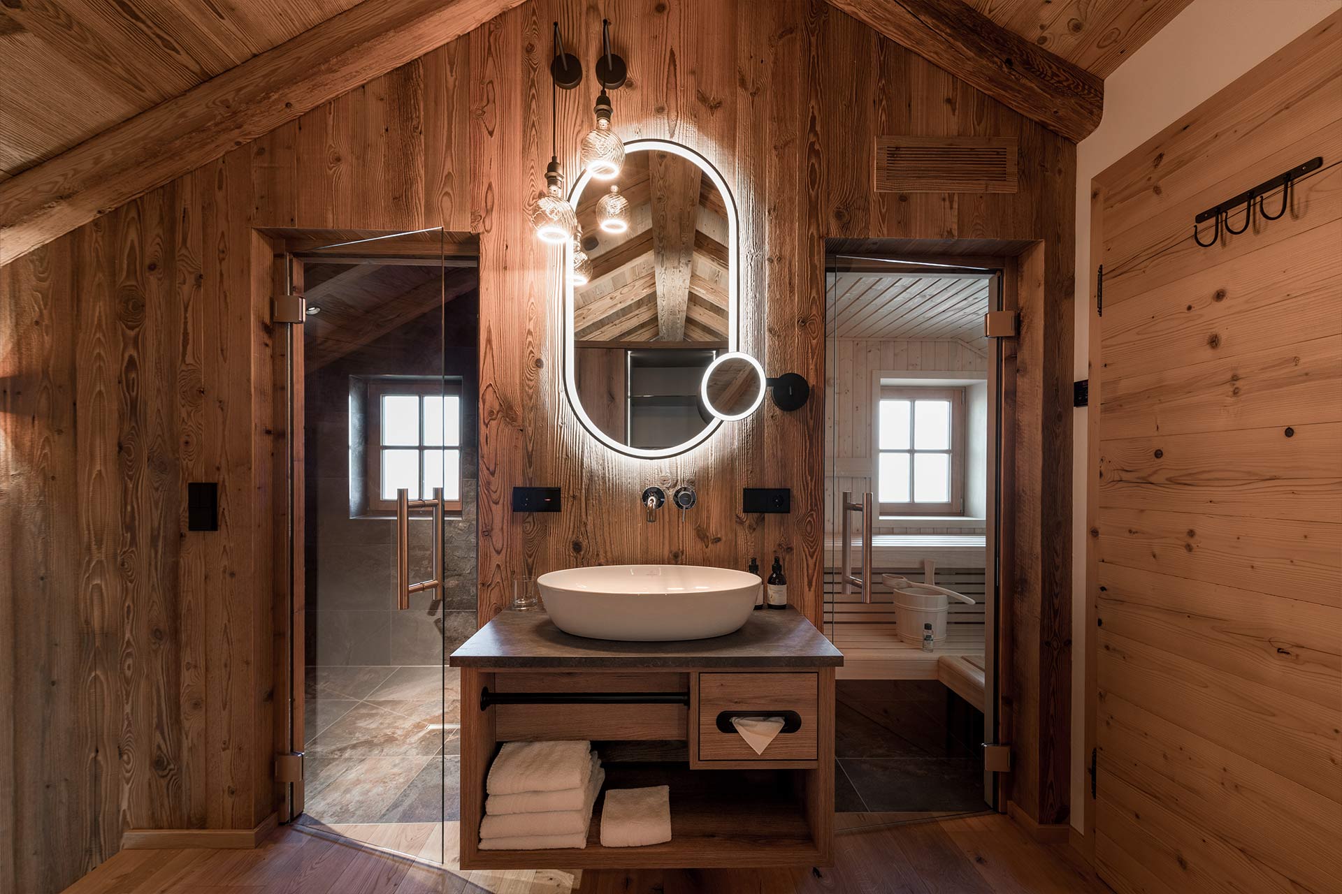 Das Bild zeigt ein stilvolles Badezimmer mit Holzverkleidung. Es gibt ein elegantes Waschbecken auf einem Holztisch, einen runden Spiegel mit Beleuchtung und große Fenster, die für natürliches Licht sorgen. Rechts ist eine Sauna sichtbar. Die Atmosphäre wirkt modern und warm.