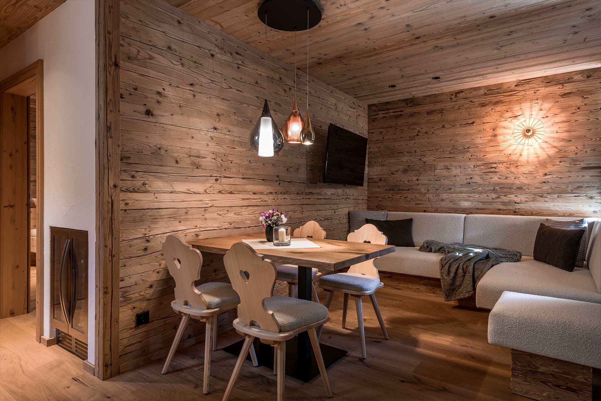 Ein gemütlicher Raum mit Holzwänden, einem Tisch und vier Stühlen. Auf dem Tisch steht eine Blumenvase. Links sieht man eine Couch mit Kissen und eine Wandlampe, die sanftes Licht ausstrahlt. Der Raum vermittelt eine warme, einladende Atmosphäre.