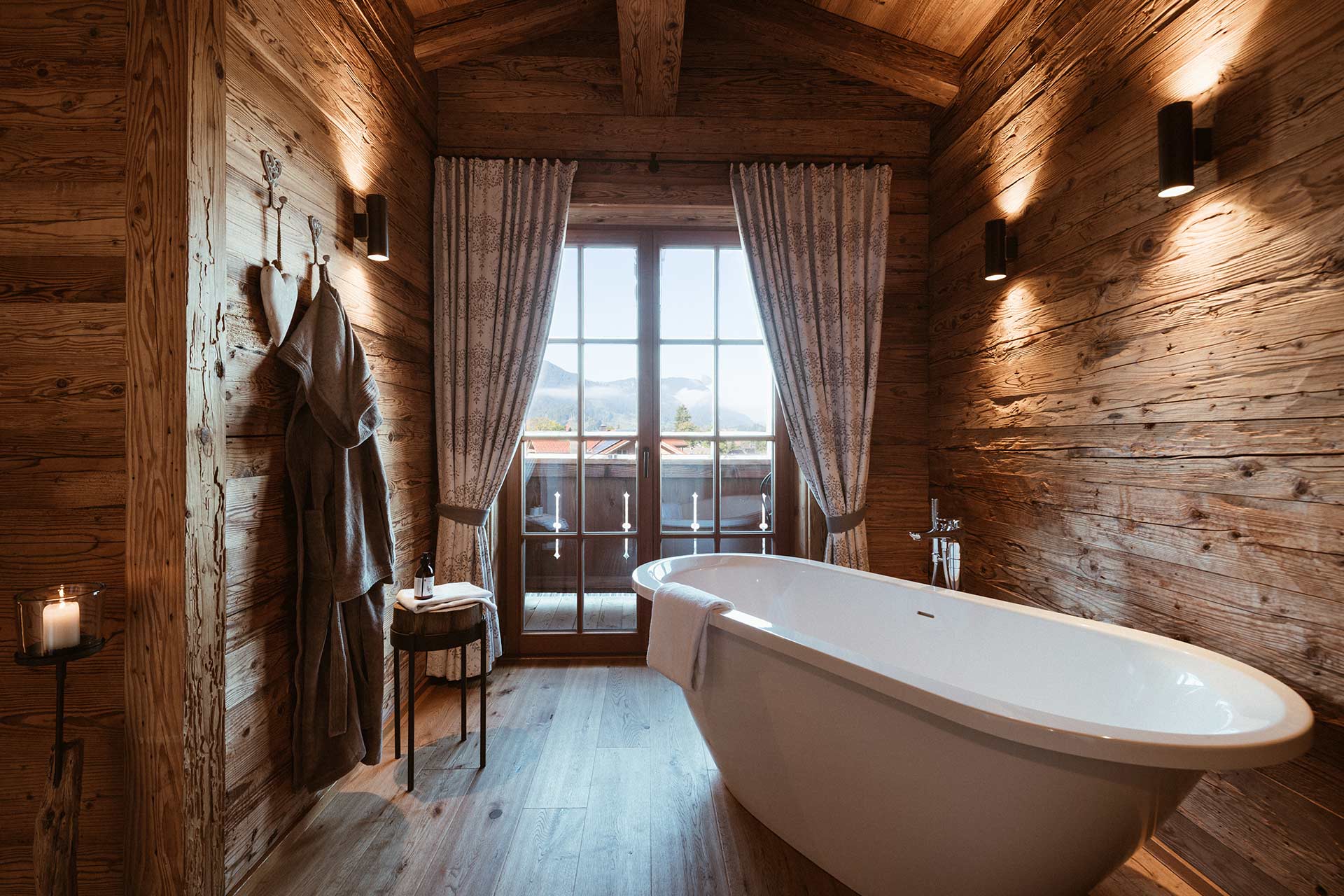 Ein modernes Badezimmer mit einer freistehenden Badewanne, Holzvertäfelung und großen Fenstern, die einen Blick auf die Landschaft bieten. Weiche Vorhänge und eine Holzbank ergänzen die gemütliche Atmosphäre. Im Hintergrund hängen zwei Bademäntel und Kerzen sorgen für stimmungsvolles Licht.