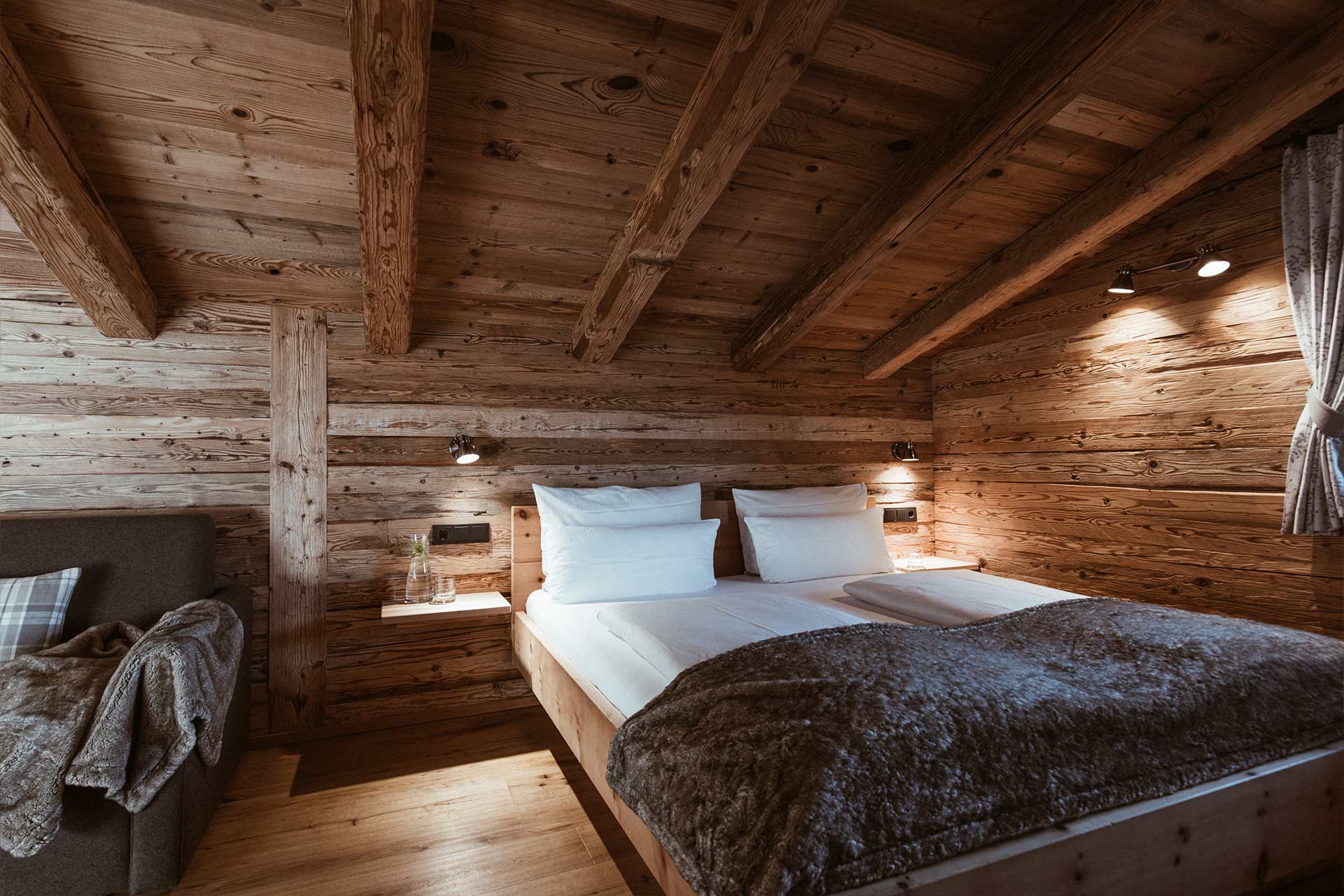 Das Bild zeigt ein gemütliches Schlafzimmer mit Holzverkleidung. Zwei Einzelbetten mit weißen Bettwäsche sind eingerichtet. Ein graues Sofa und ein warmer Überwurf sind links sichtbar. An der Wand sind zwei Lampen angebracht, und das Ambiente wirkt einladend und rustikal.