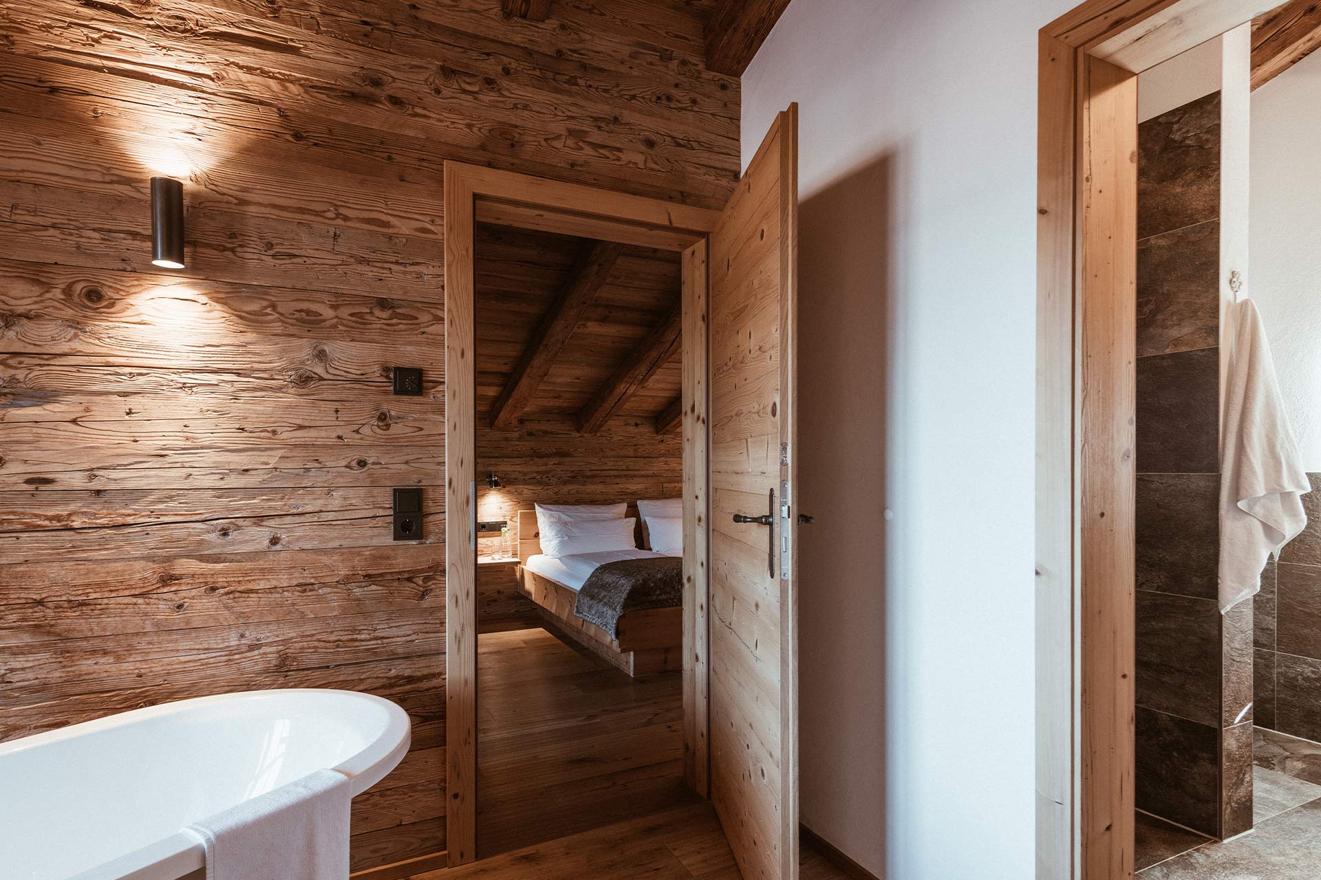Einblick in ein gemütliches, holzverkleidetes Zimmer mit einem offenen Badezimmer. Eine Badewanne mit Handtuch steht im Vordergrund, während im Hintergrund ein Bett und eine Dusche sichtbar sind. Sanftes Licht sorgt für eine warme Atmosphäre.