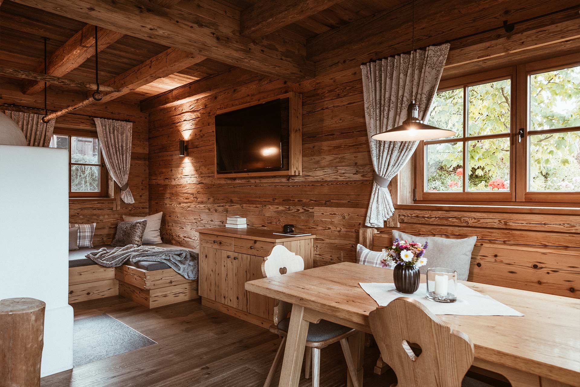 Ein rustikales Wohnzimmer in einem Holzhaus. Zu sehen sind eine gemütliche Sitzecke, ein Esstisch mit Blumen, und große Fenster, die Licht hereinlassen. Die Wände sind aus Holz, und die Fenster sind mit Vorhängen geschmückt. Ein Fernseher ist an der Wand montiert.