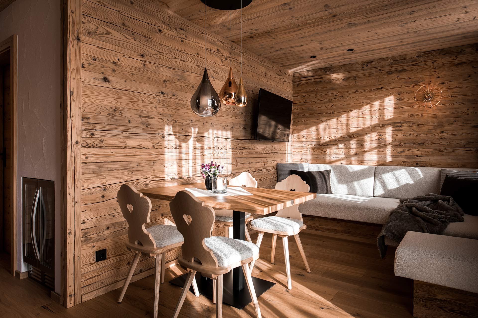 Ein gemütlicher Essbereich in einem Holzhäuschen. Ein runder Tisch mit vier Stühlen ist mit einer Blumenvase dekoriert. Im Hintergrund befindet sich eine helle Sitzgelegenheit und ein Fernseher. Weiches Licht fällt durch die Fenster und schafft eine warme Atmosphäre.