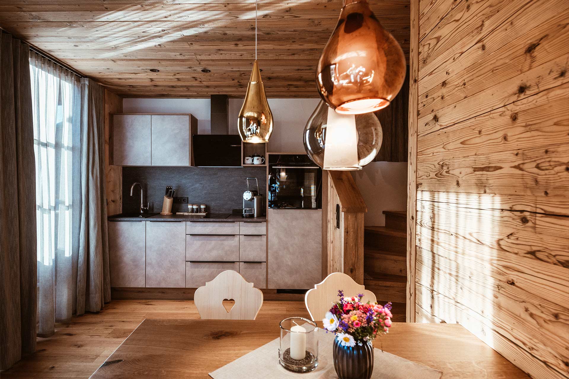 Eine helle, moderne Küche mit Holzvertäfelung. Auf dem Tisch stehen eine Kerze und ein Blumenstrauß. Über dem Tisch hängen stilvolle Lampen. Der Raum wirkt gemütlich und einladend, mit natürlichem Licht, das durch ein Fenster strömt.