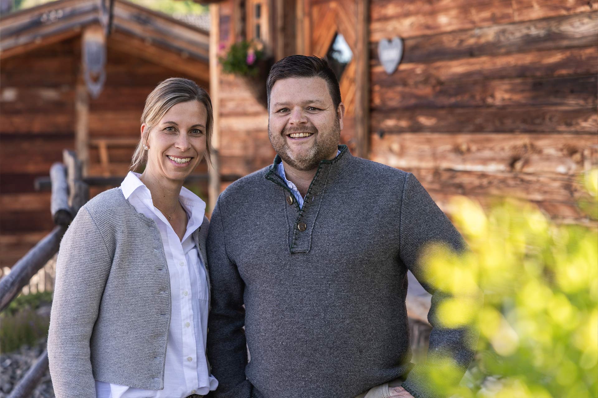 Zwei Personen stehen vor einem Holzhaus in der Natur. Die Frau trägt einen hellen Pullover über einer weißen Bluse, der Mann einen grauen Pullover. Beide lächeln in die Kamera. Im Hintergrund sind weitere Holzstrukturen und grüne Pflanzen sichtbar.