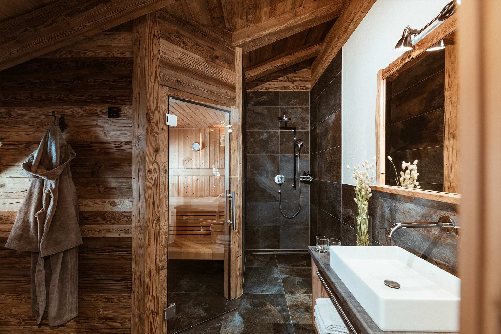 Ein gemütliches Bad mit Holzwänden und Fliesen. Auf der linken Seite ist eine Sauna mit Holzbank sichtbar. Rechts befindet sich ein Waschbecken mit modernem Wasserhahn und einer Vase mit Blumen. Ein Handtuch hängt an der Wand, und es herrscht eine entspannende Atmosphäre.