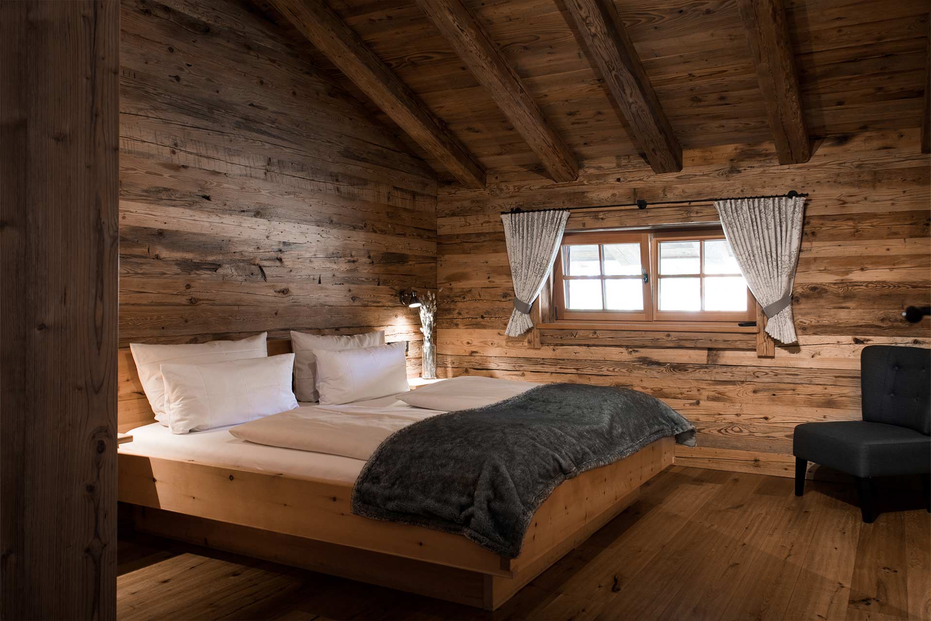 In einem gemütlichen Holzzimmer befindet sich ein Doppelbett mit frisch bezogenen Kissen und einer grauen Decke. An einem Fenster sind weiße Vorhänge befestigt. Ein bequemer Sessel steht in der Ecke, und die Wände sind aus rustikalem Holz.