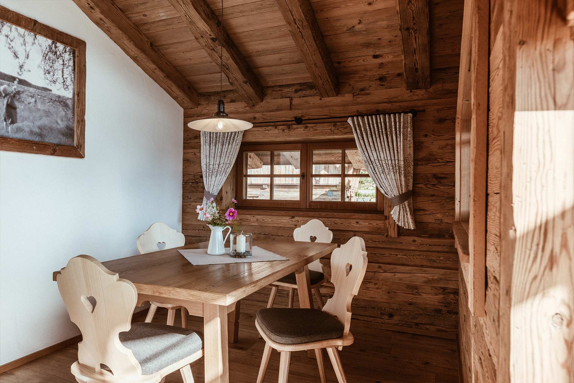 Ein gemütlicher Essbereich in einem Holzhaus. Ein Tisch mit vier Stühlen, auf dem ein Tischläufer und eine kleine Vase mit Blumen stehen. Der Raum hat große Fenster mit Vorhängen und eine Holzdecke, die eine warme Atmosphäre schafft.