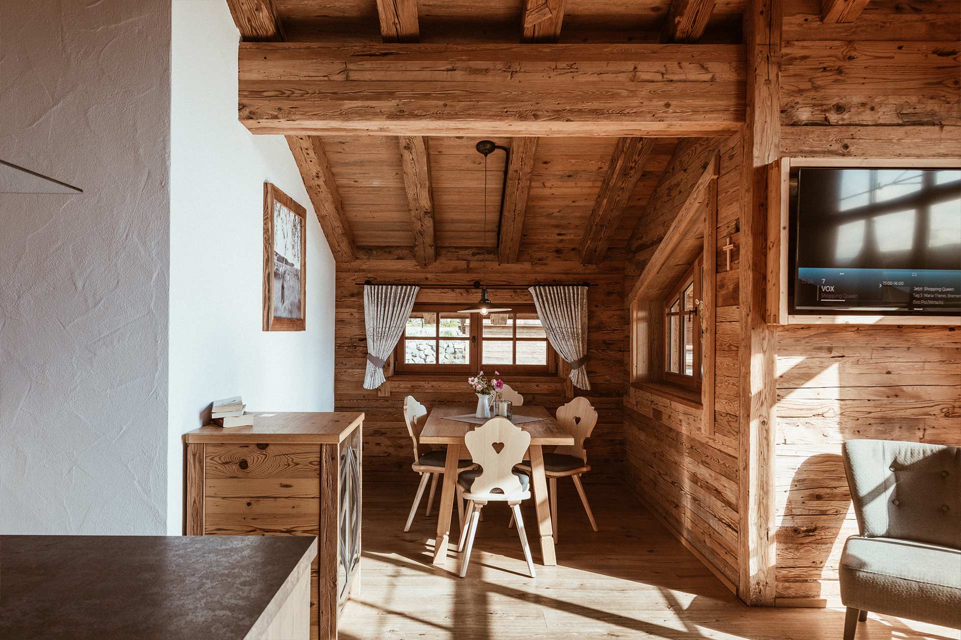 Ein helles, rustikales Esszimmer mit Holzwänden und -decken. In der Mitte steht ein Tisch mit vier Stühlen. Auf dem Tisch steht eine Vase mit Blumen. Fenster mit Vorhängen lassen Sonnenlicht herein. Ein Fernseher ist an der Wand angebracht.