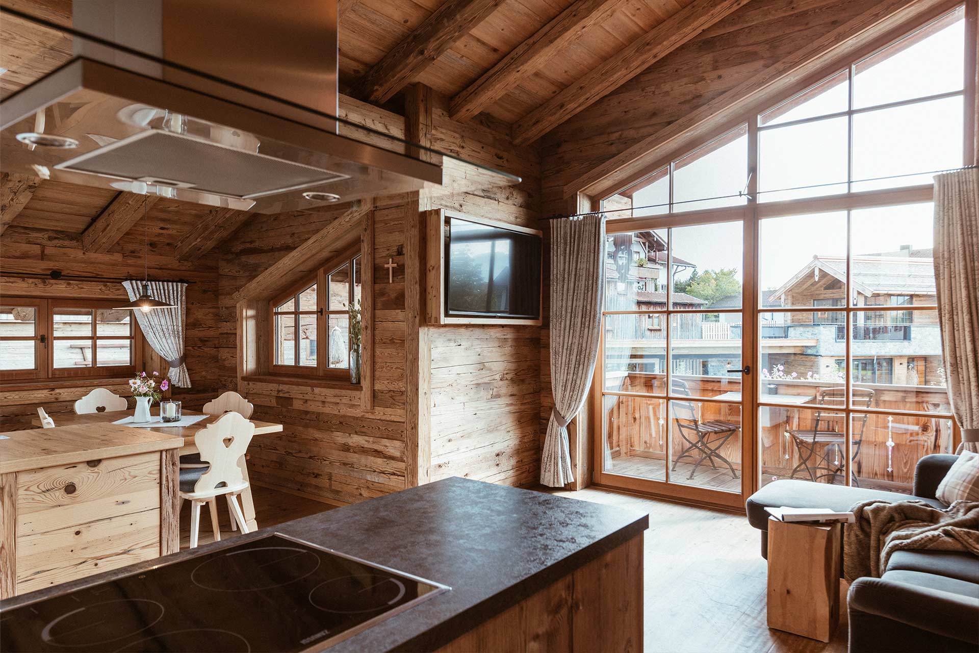 Ein gemütlicher, holzverkleideter Raum mit einer offenen Küche und einem großen Fenster, das einen Blick auf einen Balkon und die Umgebung bietet. Der Tisch ist aus Holz und hat Stühle, während eine moderne Kochinsel im Vordergrund steht.