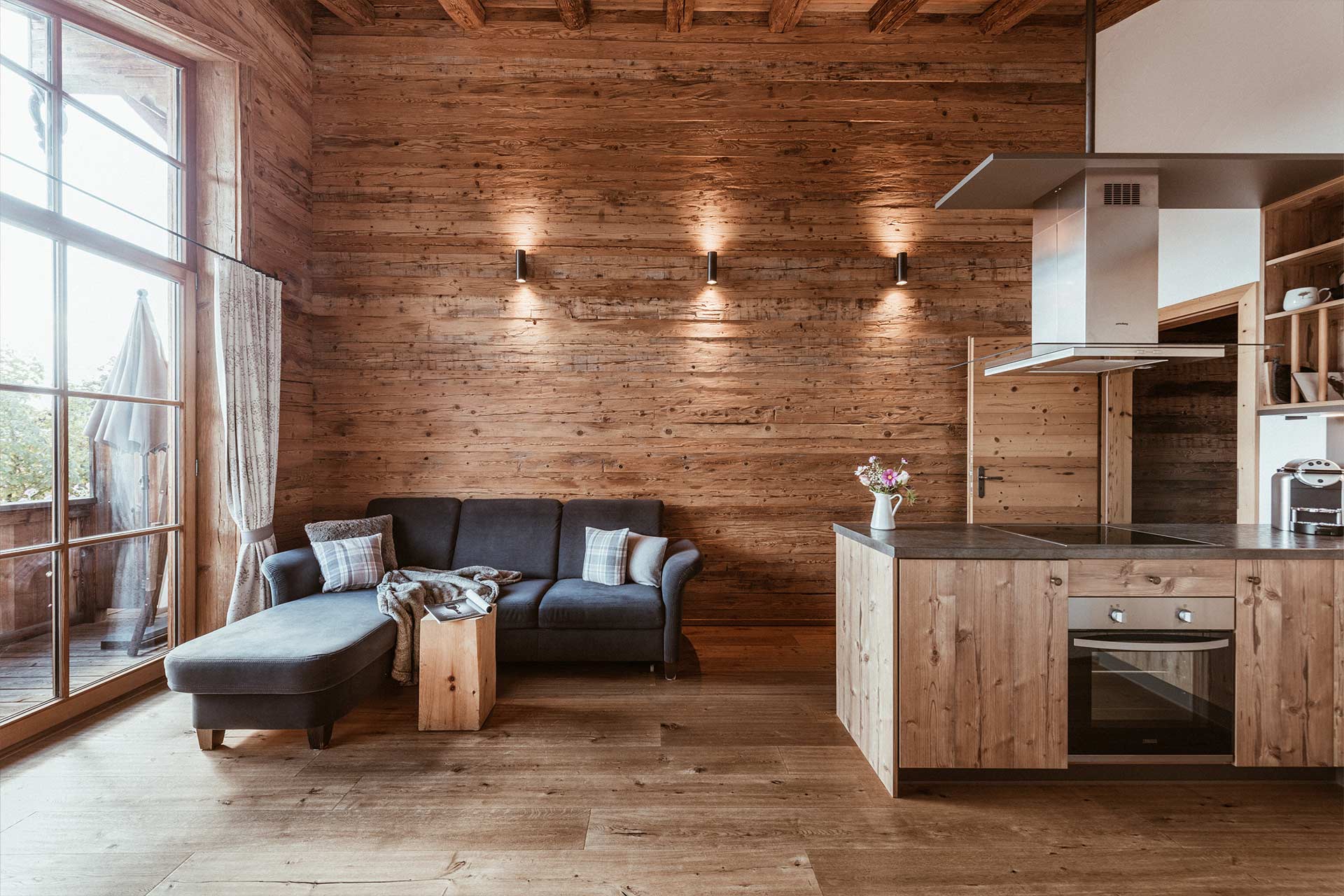 Ein modern gestalteter Wohnraum mit Holzwänden. Links befindet sich ein bequemes Sofa mit Kissen. Rechts steht eine rustikale Küchenzeile mit Holzfronten und einem Herd. Das Licht ist sanft und die Atmosphäre wirkt einladend und gemütlich.