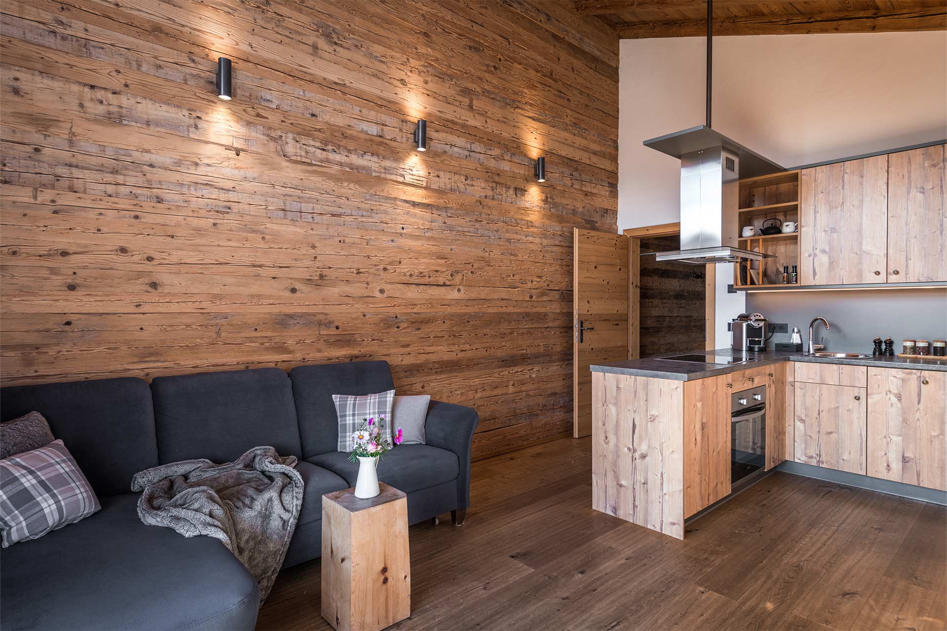 Eine moderne Holzwohnung mit einer gemütlichen grauen Couch und einem kleinen Tisch. Die Wände sind aus Holz, und die Küche ist offen mit Holzschränken und modernen Geräten. Die Beleuchtung erzeugt eine warme Atmosphäre im Raum.