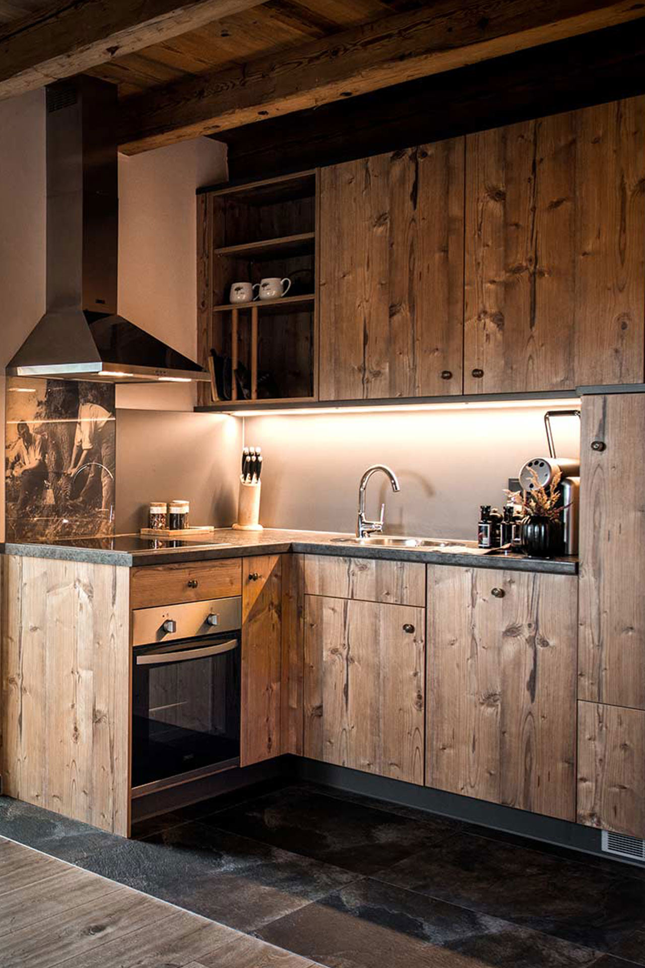 Die Bildbeschreibung zeigt eine moderne Küche mit Holzschränken. Die Küchenzeile ist mit einer Spüle, einem Herd und einer Dunstabzugshaube ausgestattet. Im Hintergrund sorgt eine sanfte Beleuchtung für eine gemütliche Atmosphäre. Küchenutensilien sind sichtbar auf der Arbeitsfläche und im Regal.