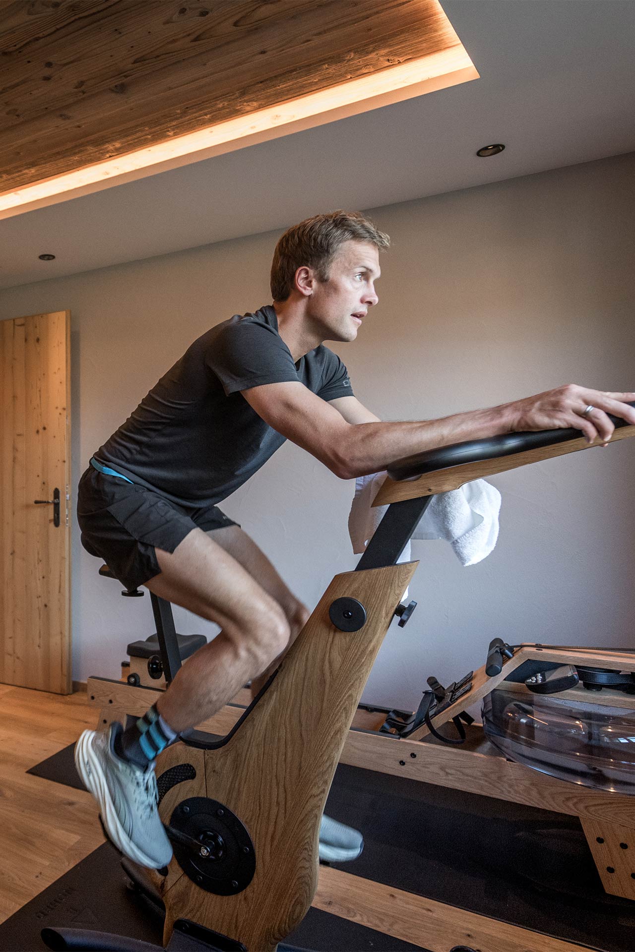 Ein Mann trainiert auf einem speziellen Fitnessgerät im Innenraum. Er sitzt auf dem Gerät, blickt konzentriert nach vorne und trägt sportliche Kleidung. Im Hintergrund sind Holz-Elemente und ein weiteres Trainingsgerät sichtbar.