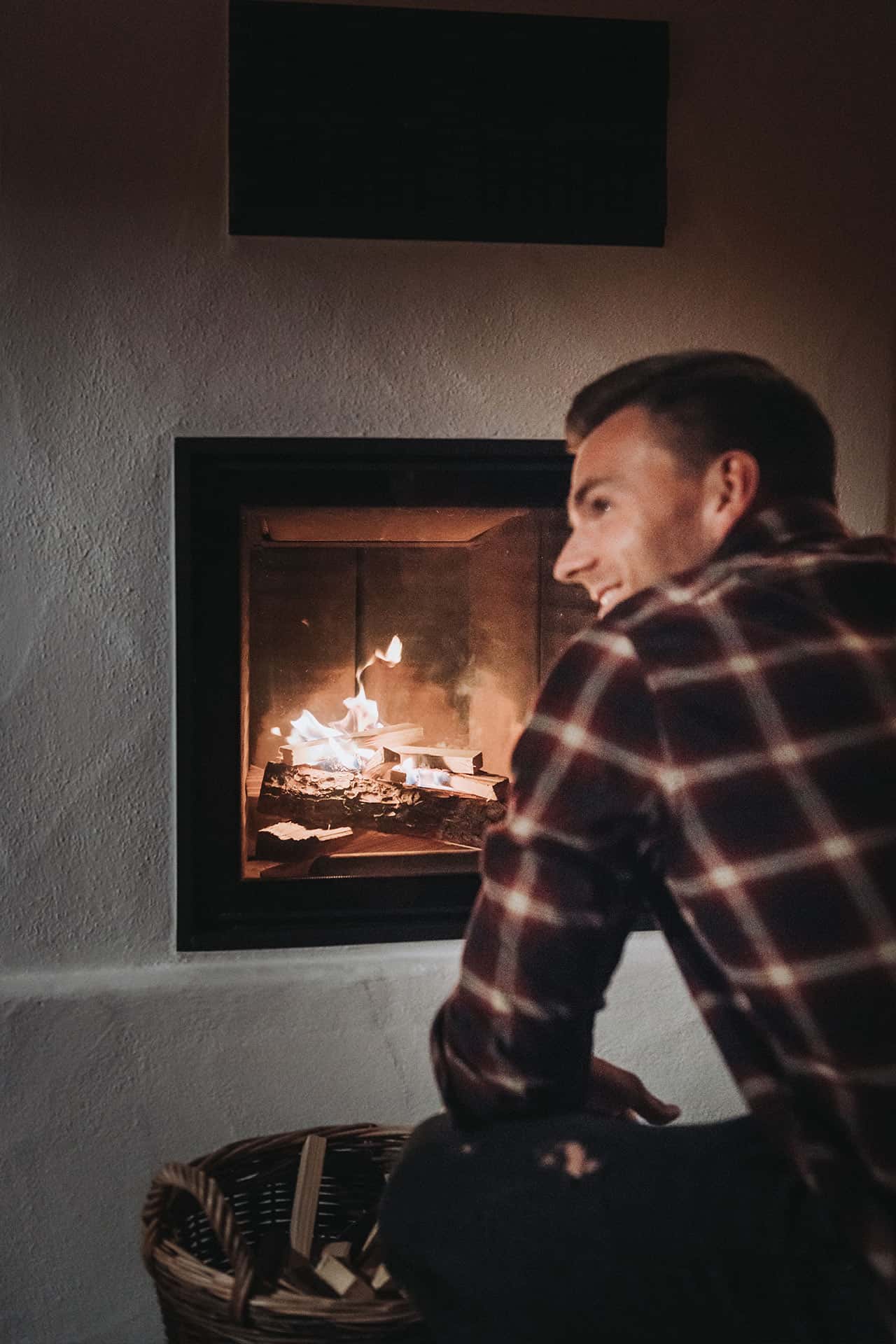 Ein Mann sitzt vor dem Kachelofen seines Chalets und wird vom frisch angezündeten Feuer beleuchtet