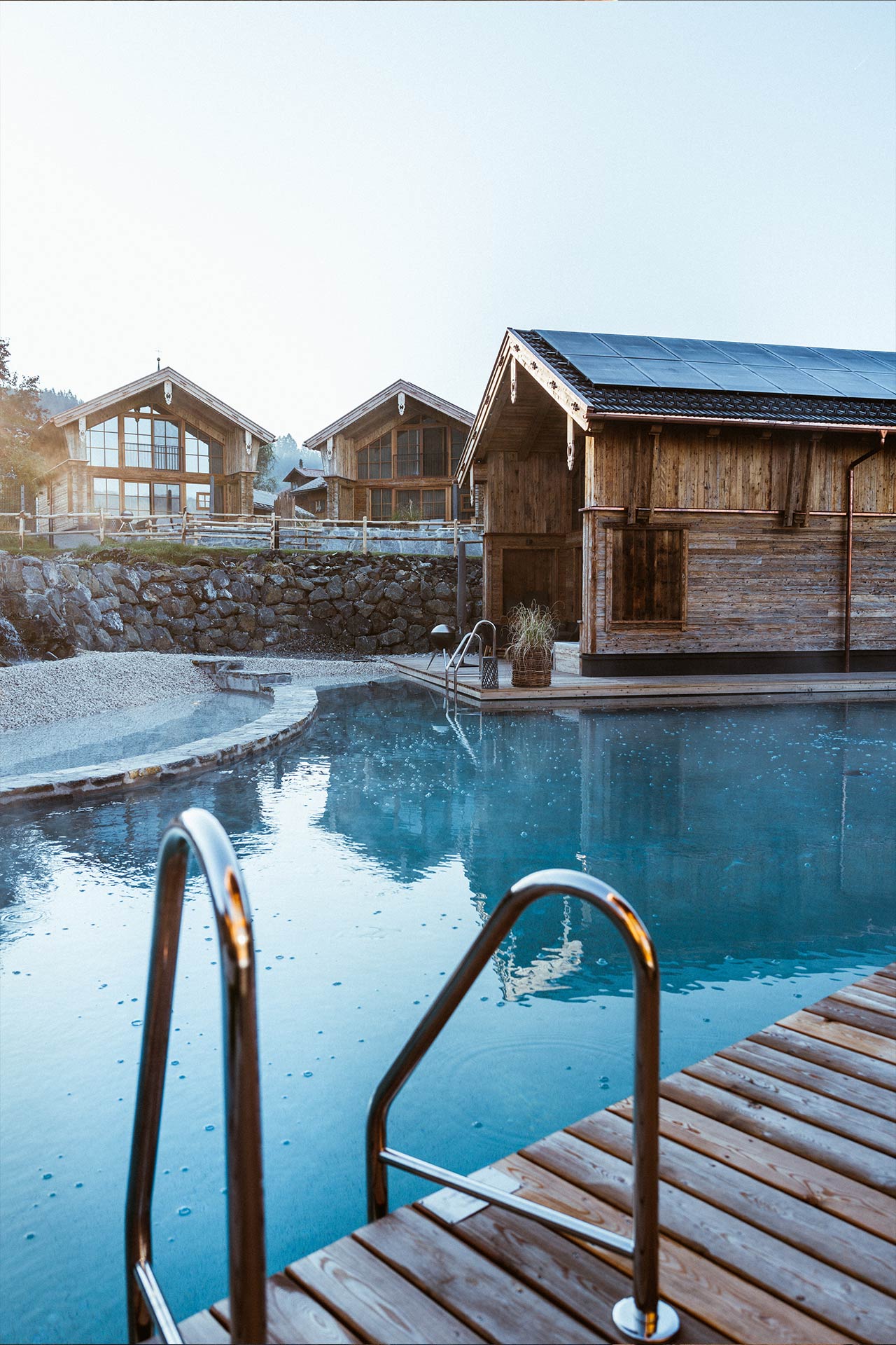 Ein ruhiger Außenpool mit einer Holzterrasse und einer Leiter. Im Hintergrund sind mehrere gemütliche Holzchalets zu sehen, umgeben von natürlicher Vegetation und sanften Hügeln. Der Himmel ist klar und leuchtet in sanften Farben.