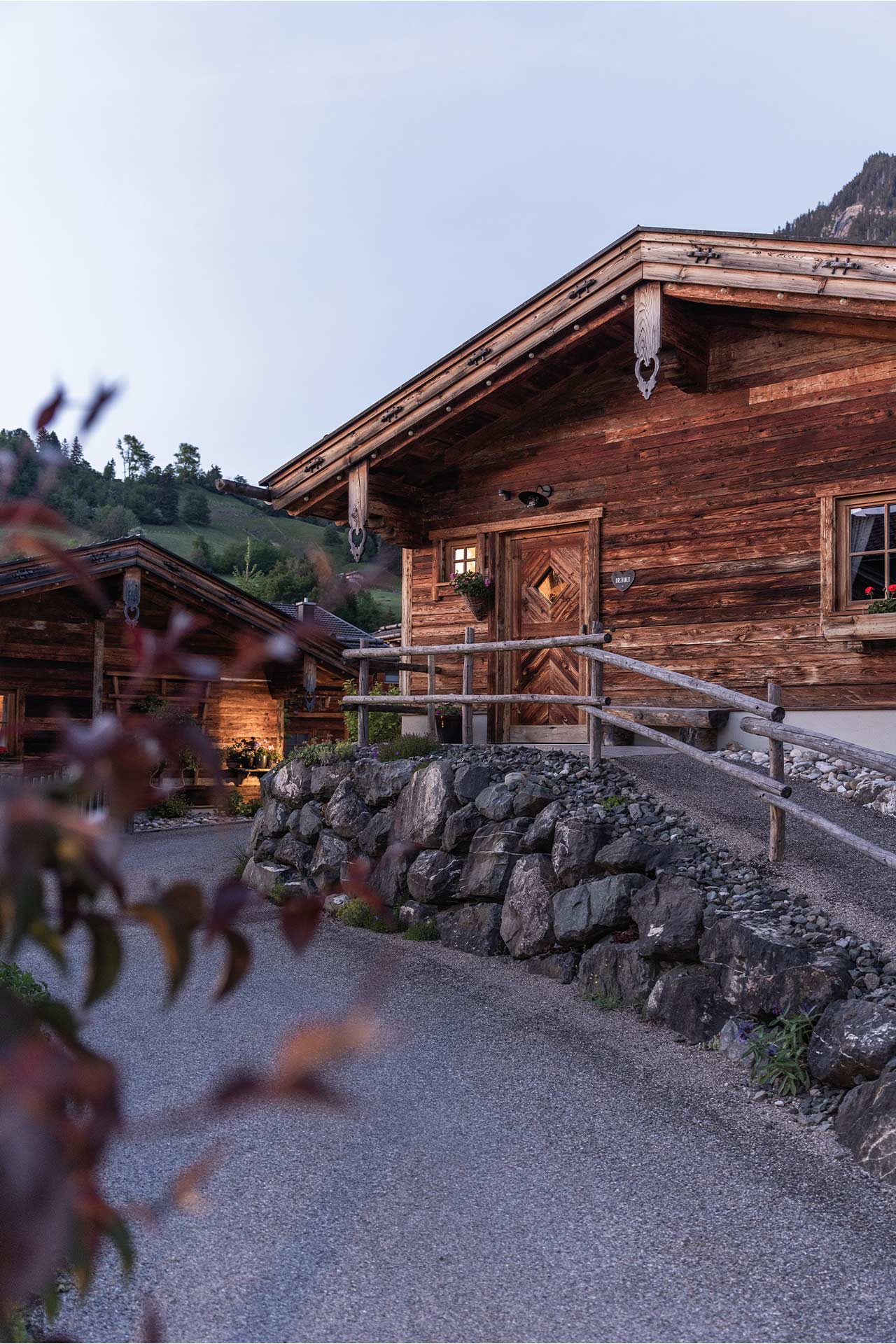 Ein traditionelles Holzhaus mit einem genauen Blick auf die Eingangstür und das Dach. Im Vordergrund ist ein Weg aus Kies zu sehen, der an einer Steinmauer entlangführt. Die Umgebung ist ländlich mit sanften Hügeln im Hintergrund und leicht gedämpftem Licht.